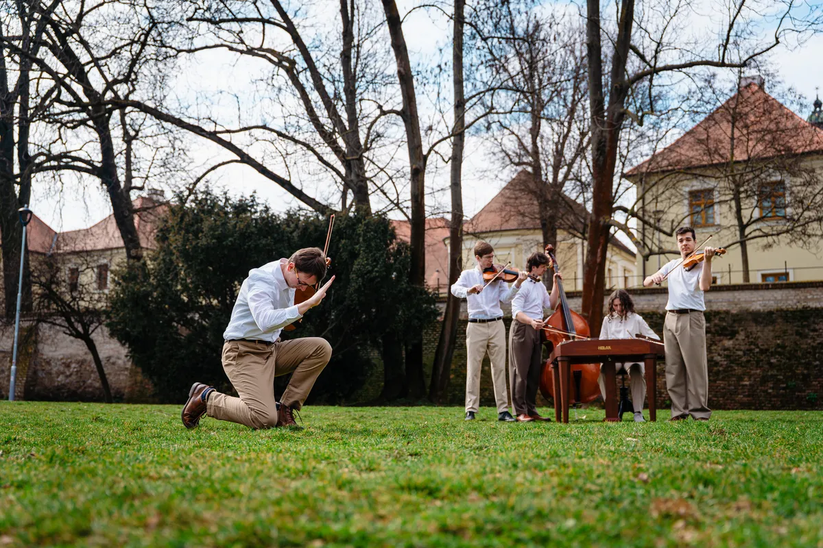 Kapela hraje na trávě — houslista v popředí, ostatní muzikanti s cimbálem a kontrabasem v pozadí